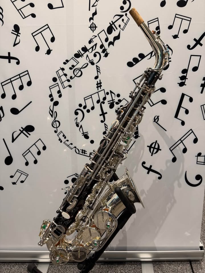 A silver alto saxophone with a light wood mouthpiece and small green crystal accents, displayed on a stand against a white wall decorated with black musical notes.